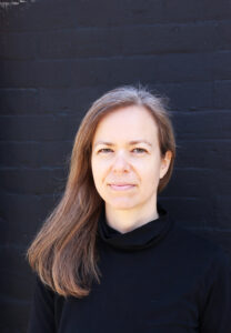 a headshot with black background. Sofya is wearing a black turtleneck. She is a white person in her 40s with long brown hair.